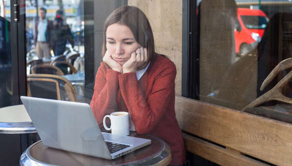 Woman sitting at table with her laptop computer and cup of coffee, resting her head in her hands