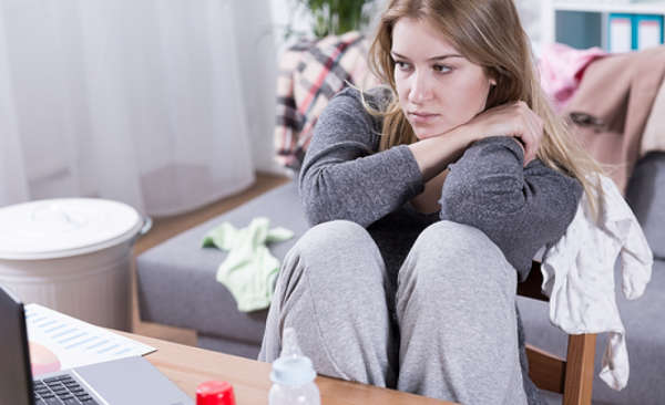 Young woman sitting in chair with diapers and baby clothes strewn about and looking stressed