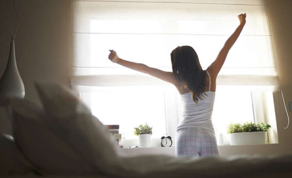 Young woman, standing by bed in bedroom, stretching after just having woken up